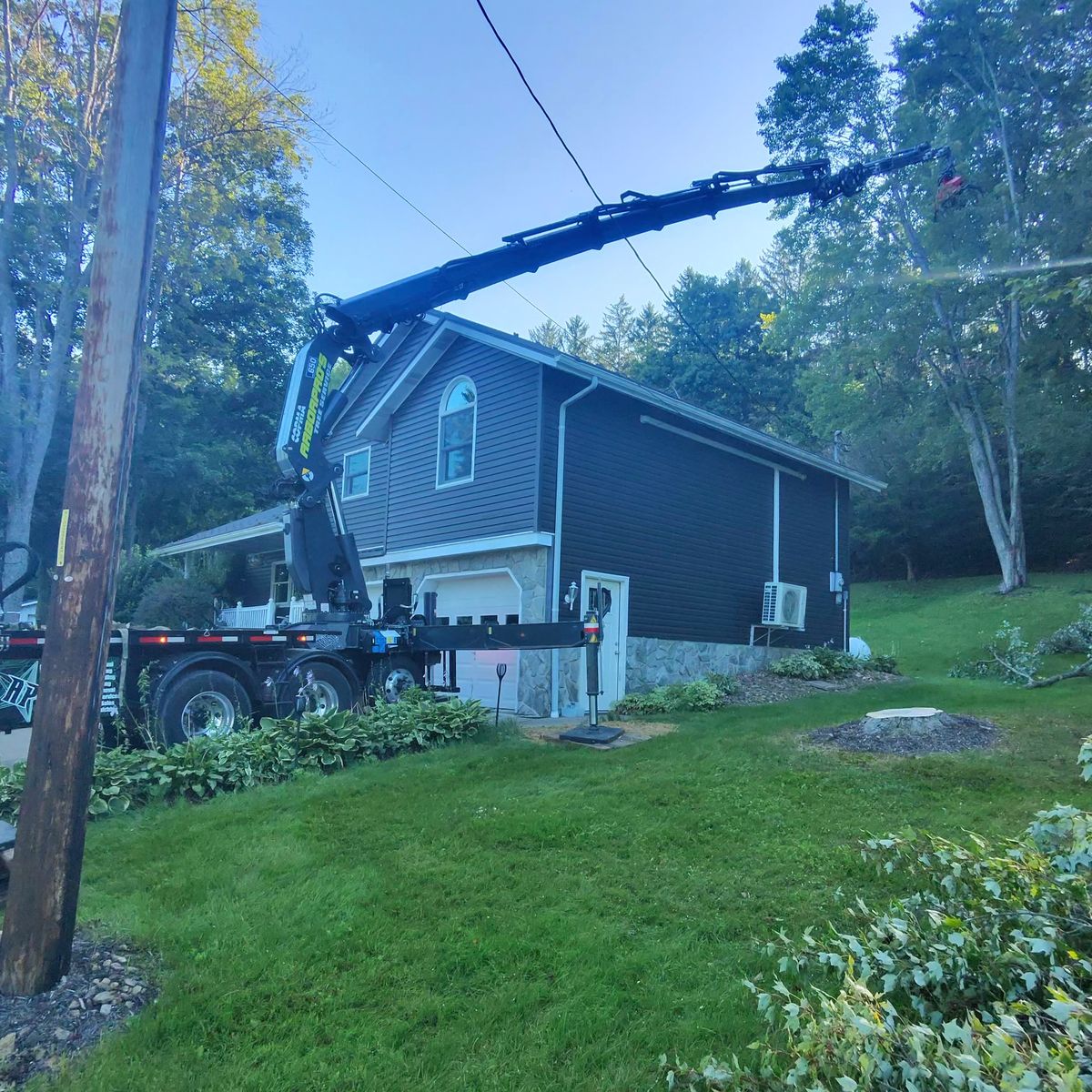 Crane reaching over a gray house to lift a tree section